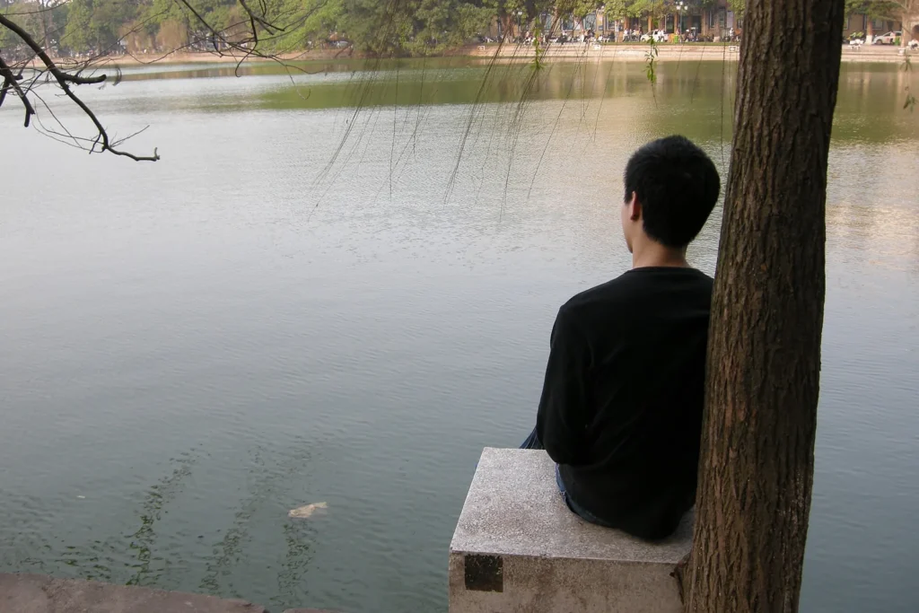 A man sitting on a stone bench under a tree beside Hoan Kiem Lake in Hanoi