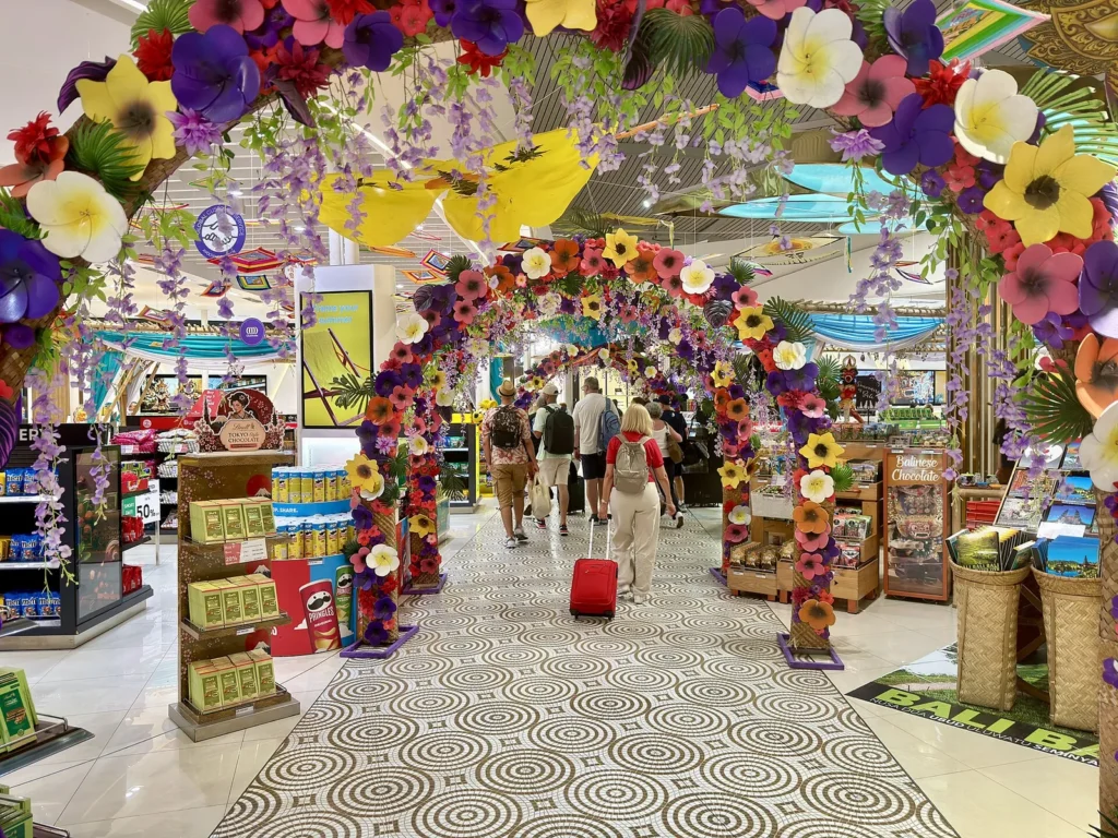 Travellers walking under a large arch of colourful paper flowers between shops at Ngurah Rai airport